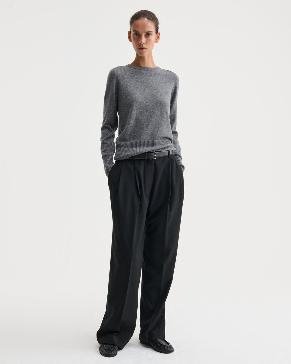 A model stands against a light gray studio background wearing a gray knit sweater, black wide-leg trousers, a black belt, and black shoes.