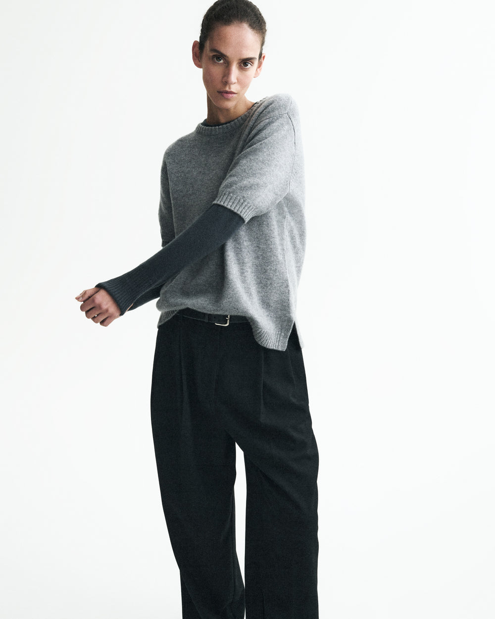 A model wearing a light gray knit sweater with long dark sleeves and black trousers, standing against a plain white studio background.