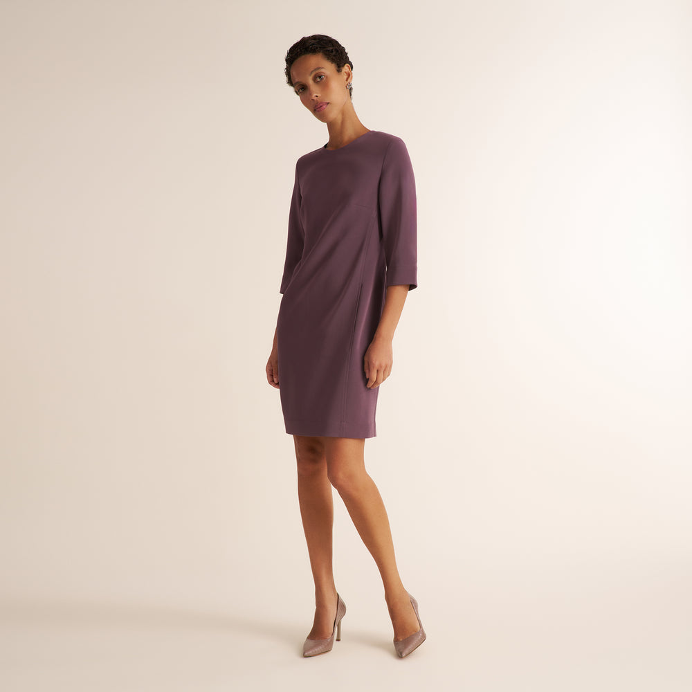 A woman modeling a knee-length, three-quarter sleeve dress in a muted plum purple color, paired with nude high heels, against a solid light beige studio background.