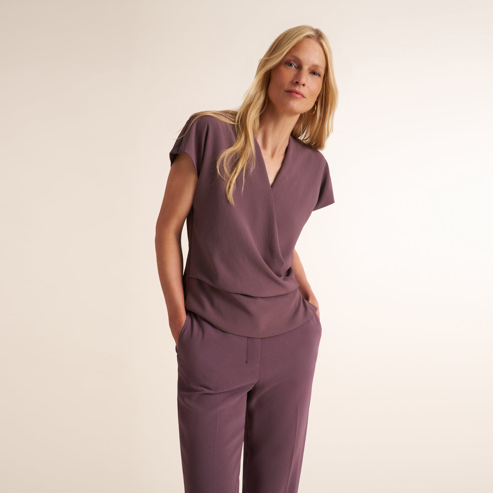 A woman modeling a matching mauve-purple scrub top and scrub pants set against a plain, warm off-white studio background.