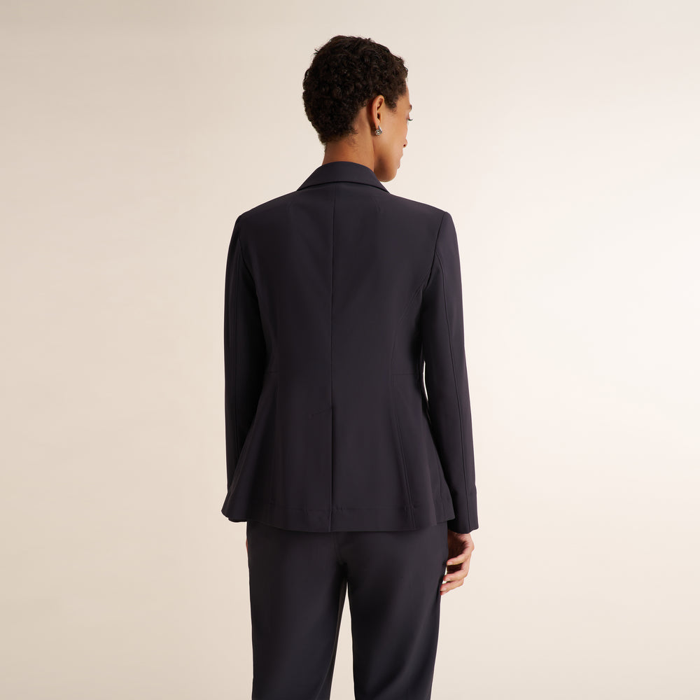 A woman modeling a matching charcoal pantsuit with a blazer and straight-leg trousers, styled with a light blue top and brown strappy heels against a plain beige studio