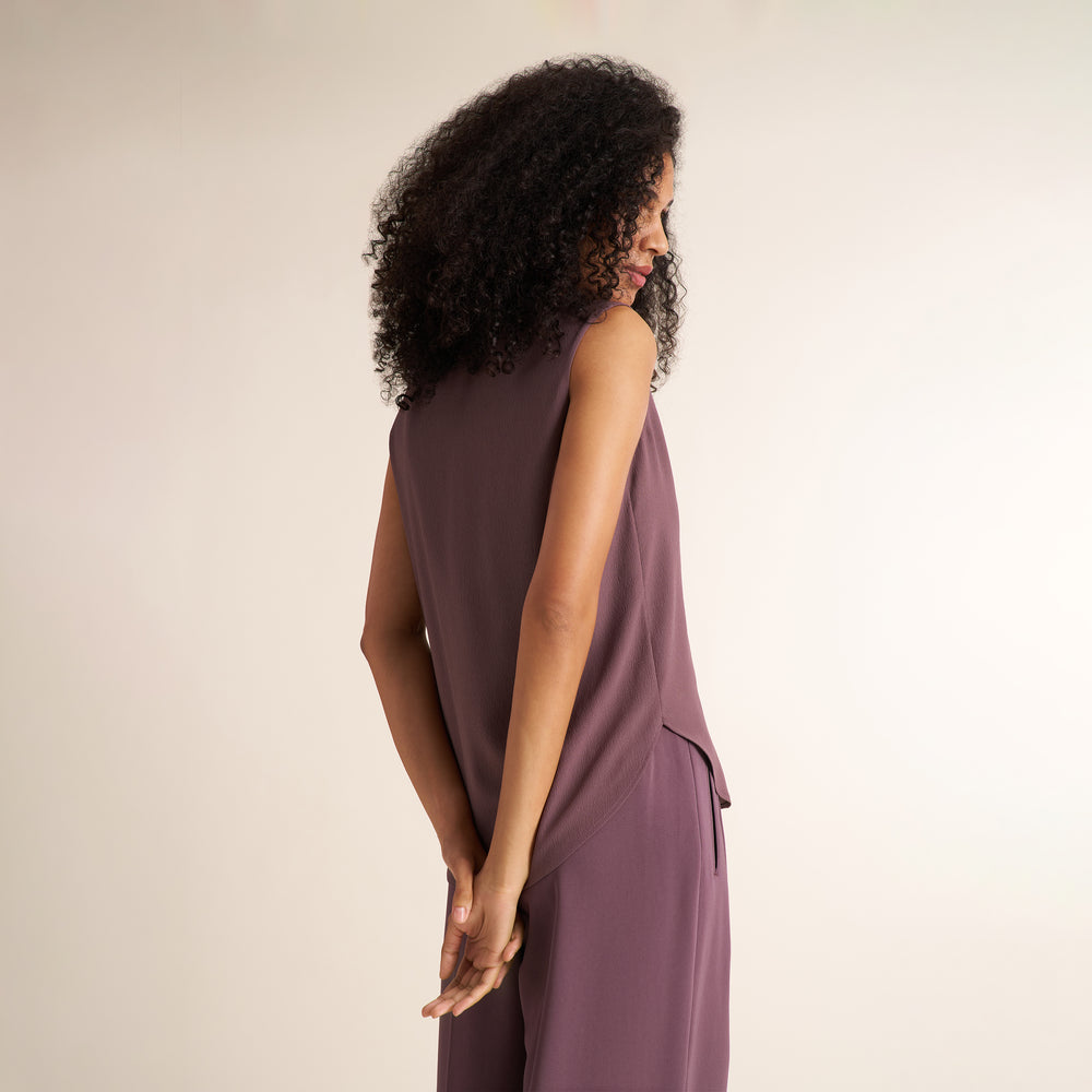 A woman viewed from behind wearing a sleeveless mauve-purple jumpsuit with a matching tie belt and wide-leg trousers, set against a plain beige studio background.