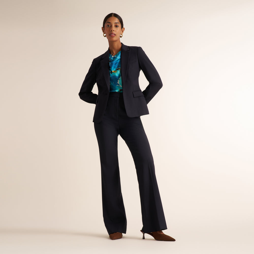 A model stands in a studio wearing a dark navy blue tailored pantsuit with a structured blazer and wide-leg trousers, styled with a colorful blue and green patterned top and brown pointed-toe heels against a warm beige background.