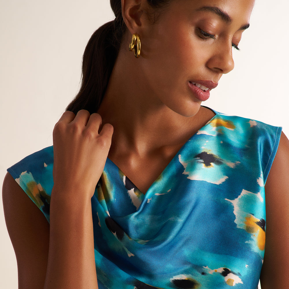 A close-up of a woman wearing a blue satin-like top with an abstract watercolor pattern in teal, yellow, and black, paired with gold hoop earrings against a plain beige background.
