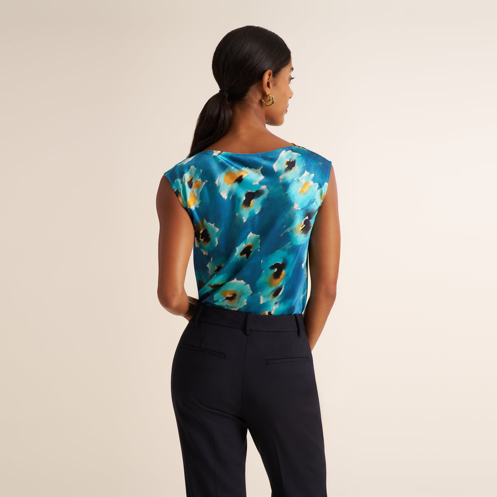 A woman shown from the back wearing a sleeveless top with a blue abstract floral print and dark navy trousers, photographed against a plain light beige studio background.