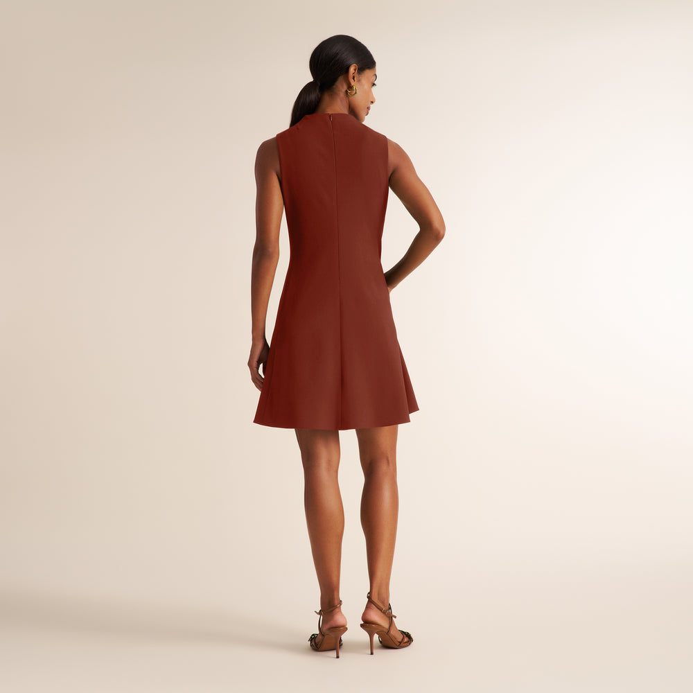 A woman is shown from the back wearing a sleeveless rust-brown shift dress and brown strappy high-heeled sandals against a plain beige studio background.