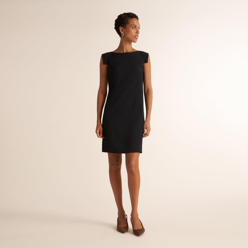 A woman modeling a black sleeveless shift dress with a boat neckline, photographed against a plain warm off-white studio background.