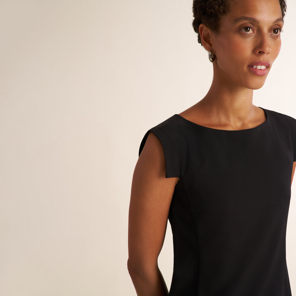A woman wearing a black sleeveless top with a boat neckline, shown from the chest up against a plain light beige studio background.