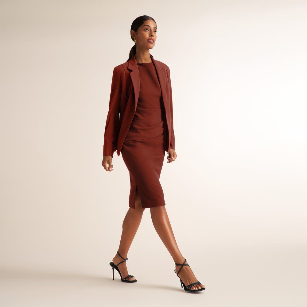 A woman models a matching rust-brown dress and blazer set with black strappy heels against a plain, warm off-white studio background.