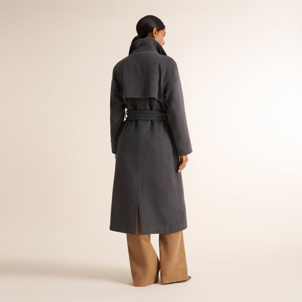 A model shown from the back wearing a dark gray belted coat over tan wide-leg trousers, photographed against a plain light beige studio background.