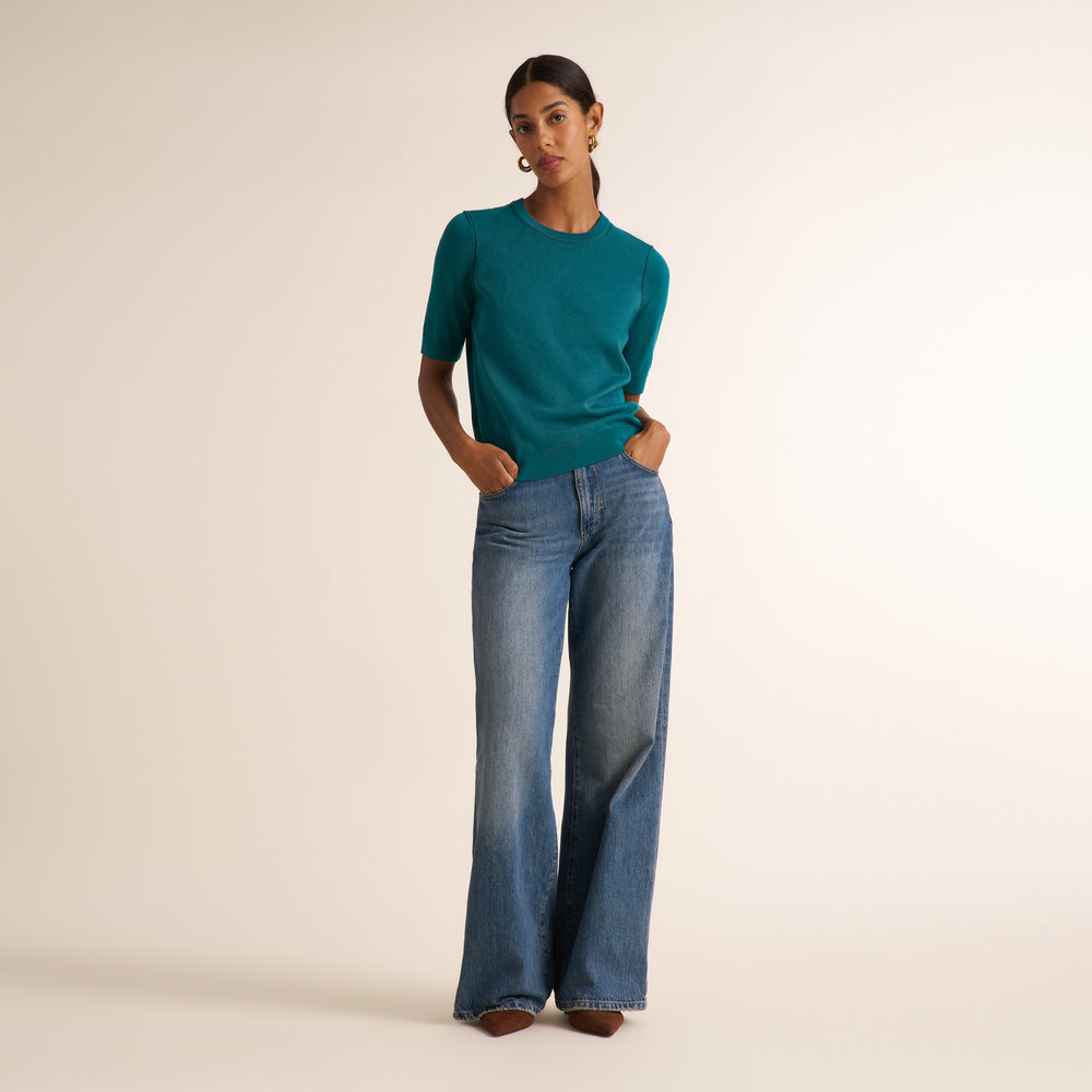 A woman modeling a teal short-sleeve knit top and wide-leg blue jeans against a plain beige studio background.