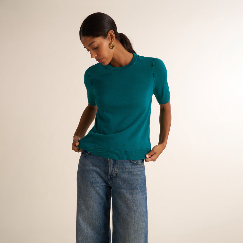 A woman wearing a teal short-sleeve knit top and blue jeans, standing against a plain light beige studio background.