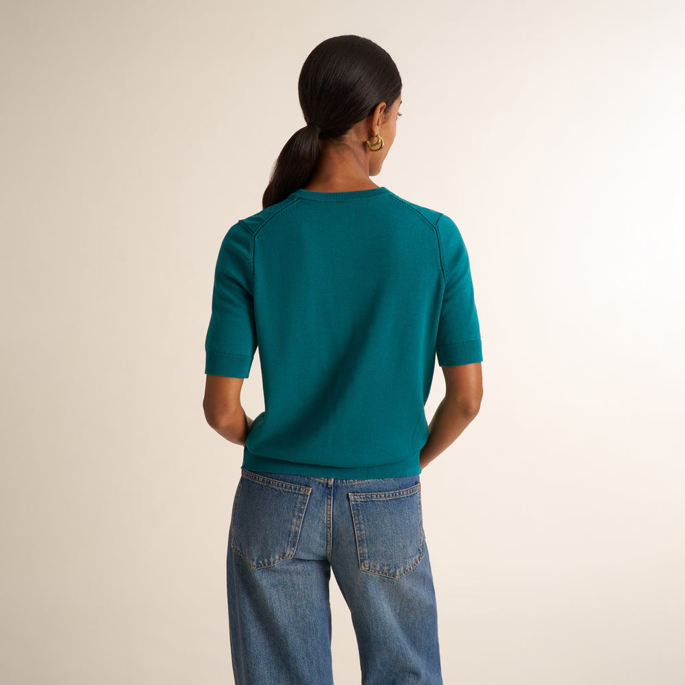 A woman viewed from behind wearing a teal short-sleeve knit top and blue jeans against a plain light beige studio background.