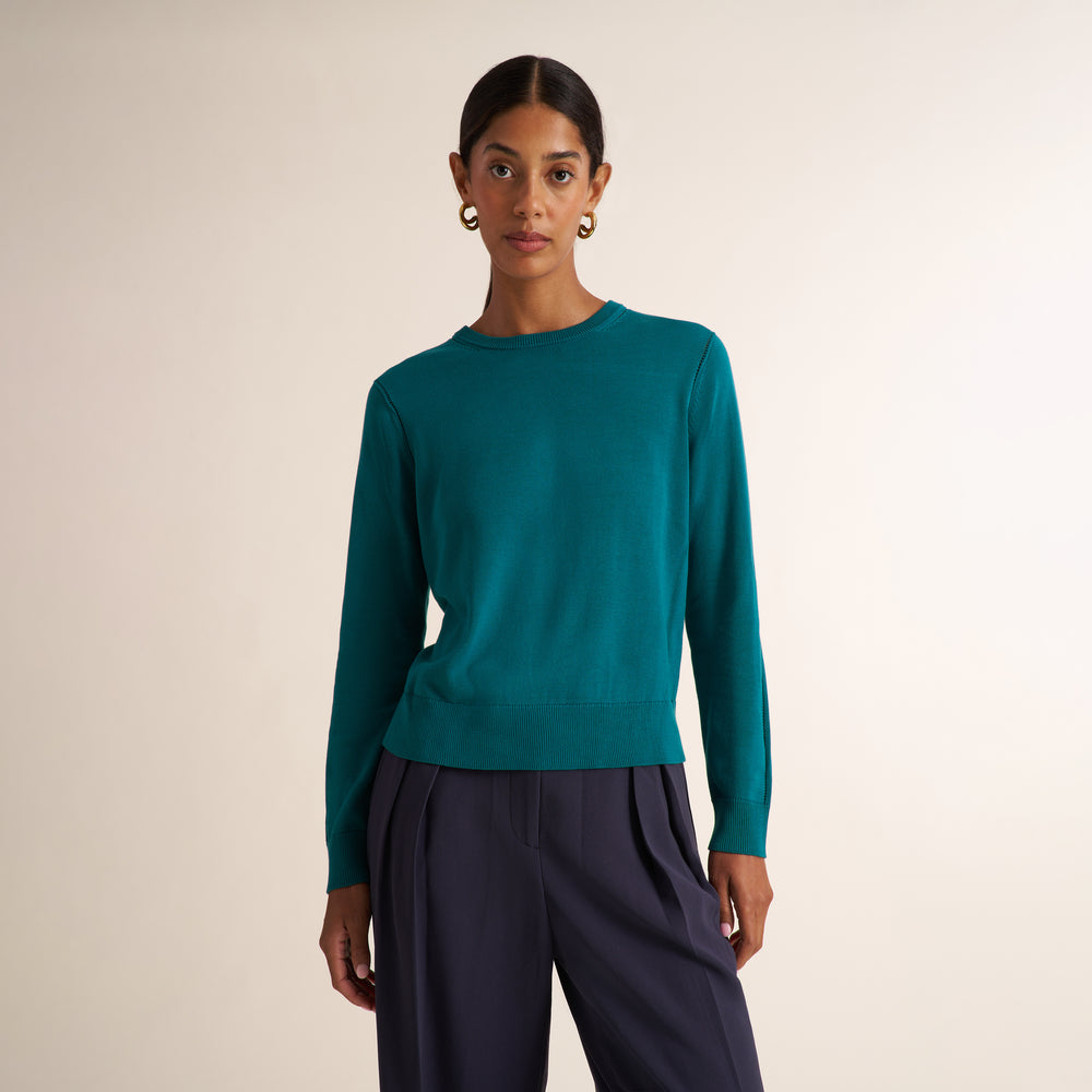 A woman modeling a teal long-sleeve knit sweater with dark navy wide-leg trousers, standing against a plain beige studio background.