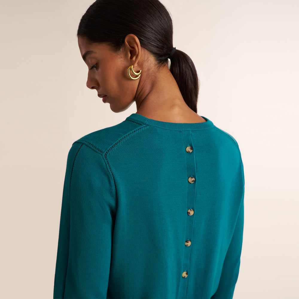 A woman shown from the back wearing a teal long-sleeve knit top with a vertical row of gold buttons down the center. She is also wearing gold hoop earrings, set against a plain beige studio background.