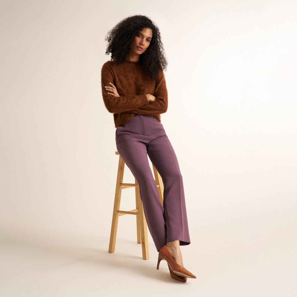 A model with curly dark hair sits on a wooden stool, wearing a brown textured knit sweater, mauve-purple wide-leg trousers, and brown pointed-toe heels against a warm off-white studio background.