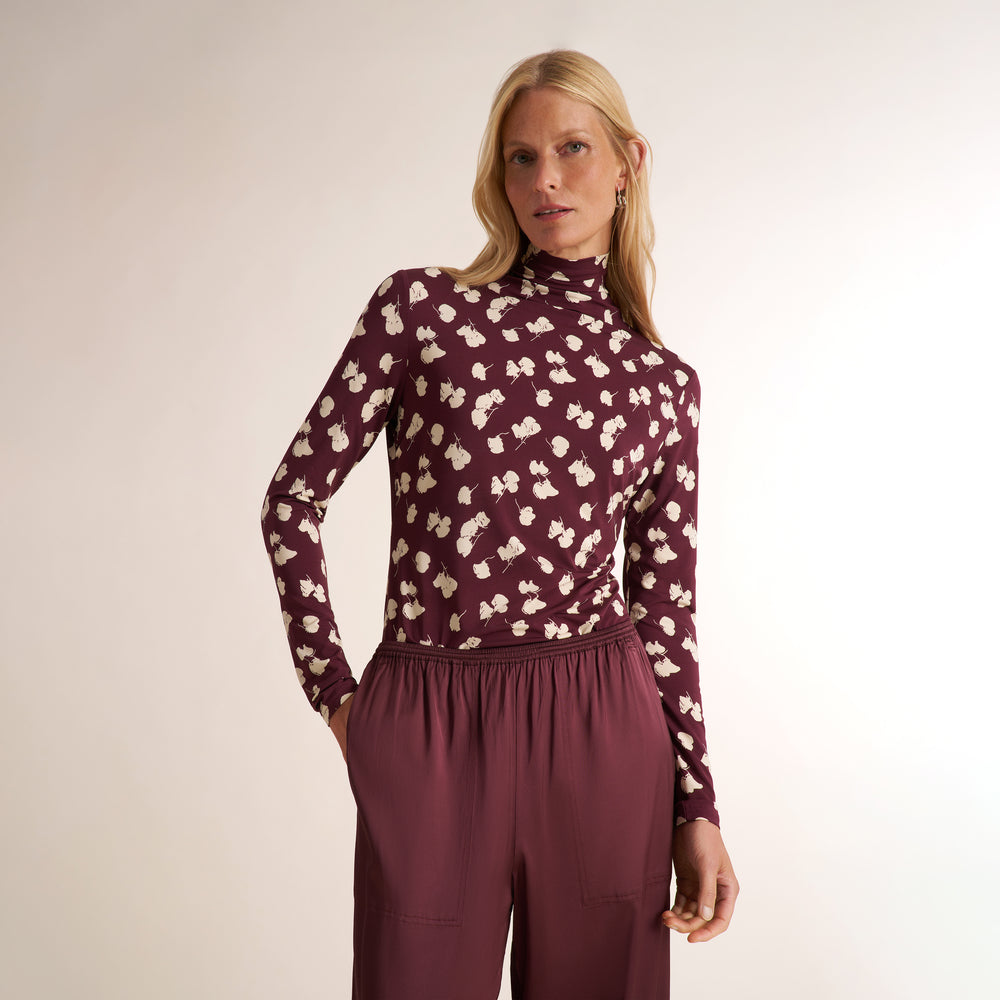 A woman modeling a long-sleeve mock neck top in burgundy with a cream abstract floral pattern, paired with matching burgundy drawstring trousers, against a plain light beige studio background.