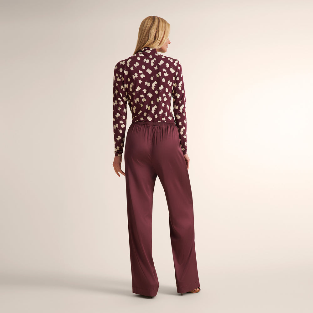A woman is shown from behind wearing a long-sleeve burgundy blouse with a white floral pattern and matching wide-leg trousers against a plain studio background.