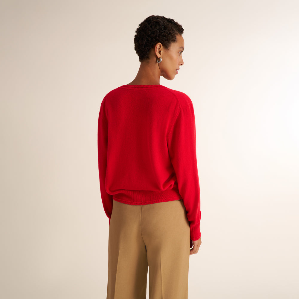 A woman shown from the back wearing a bright red knit sweater and beige wide-leg trousers, photographed against a plain light beige studio background.