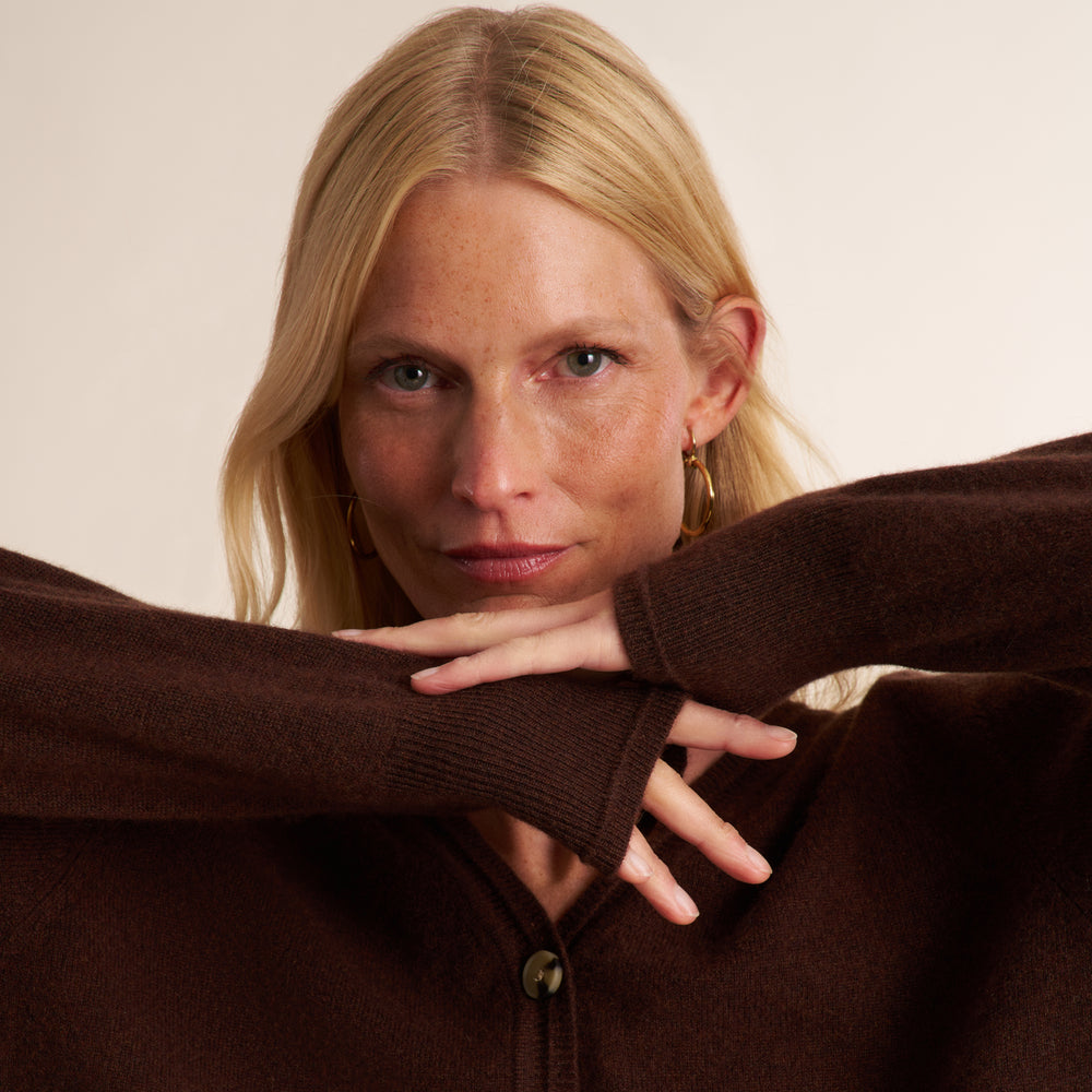 A close-up portrait of a blonde woman wearing a dark brown knit cardigan sweater, with her arms crossed over her chest against a plain light beige background.