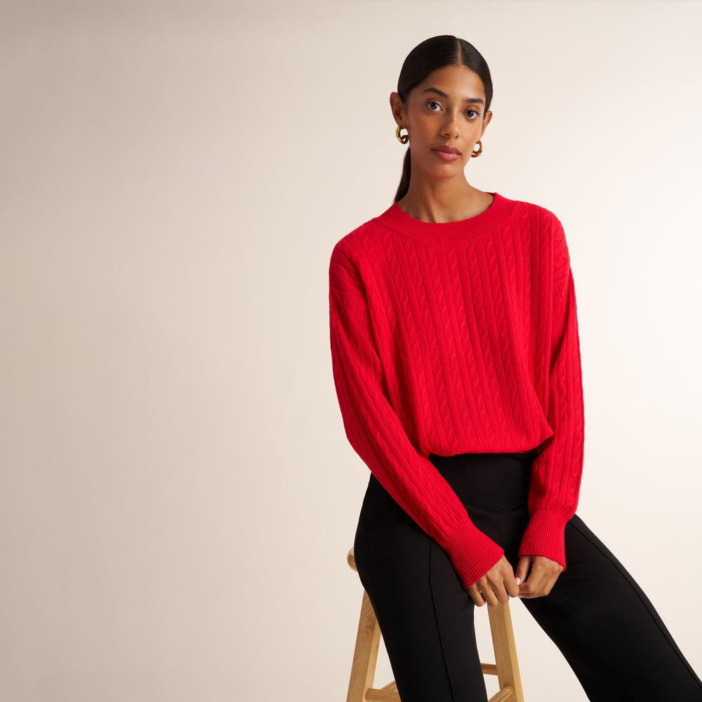 A woman wearing a bright red cable-knit long-sleeve sweater and black pants, seated on a wooden stool against a plain light beige studio background.