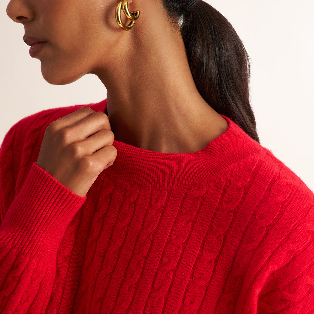 Close-up of a person wearing a bright red cable-knit crewneck sweater, with a gold hoop earring visible against a plain light background.