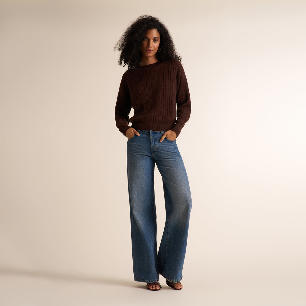 A woman modeling a dark brown cable-knit sweater and wide-leg blue jeans against a plain beige studio background.