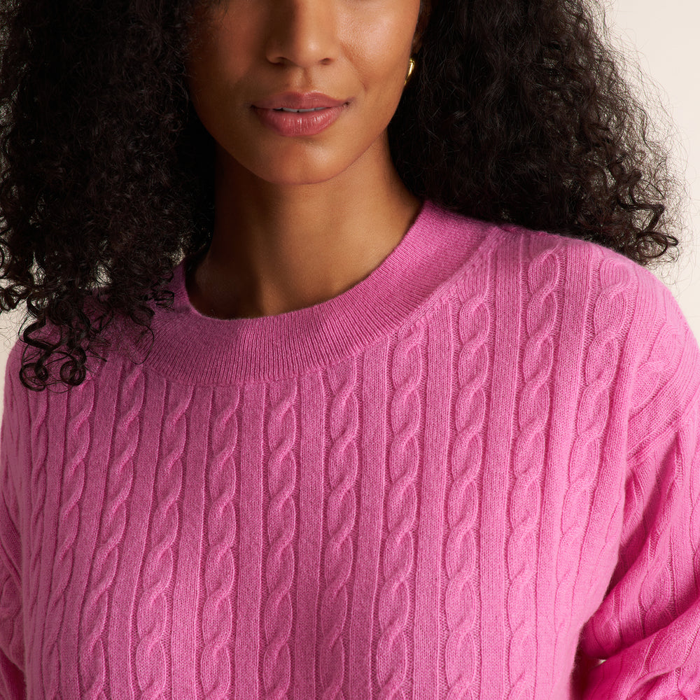 A close-up of a woman wearing a bright pink cable-knit crewneck sweater, showing the detailed knit texture and her dark curly hair against a light neutral background.