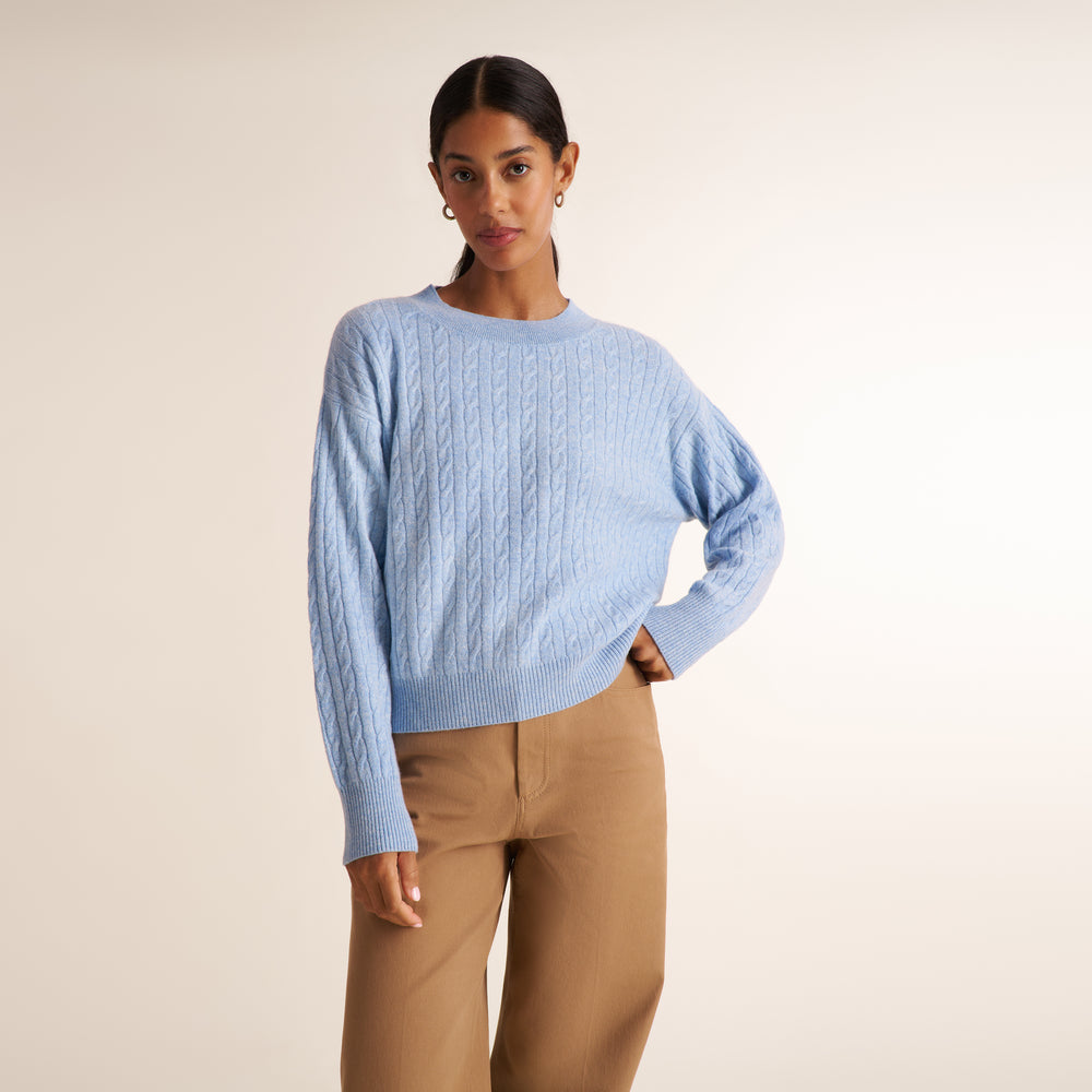 A brunette woman modeling a blue melange long-sleeve knit top with a a crew neck collar and cable knit detail, paired with saddle wide leg trousers, against a plain light beige studio background.