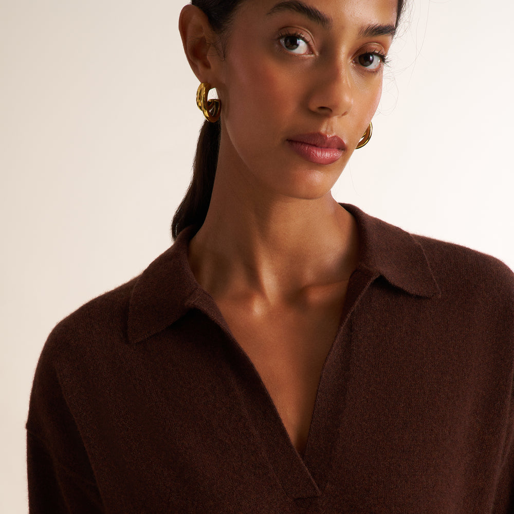 A close-up of a woman wearing a dark brown knit collared sweater and gold hoop earrings against a plain light background.