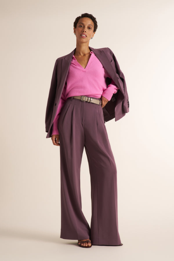 A brunette woman modeling a pink long-sleeve knit top with a split V-neck collar, paired with purple trousers and purple blazer, against a plain light beige studio background.