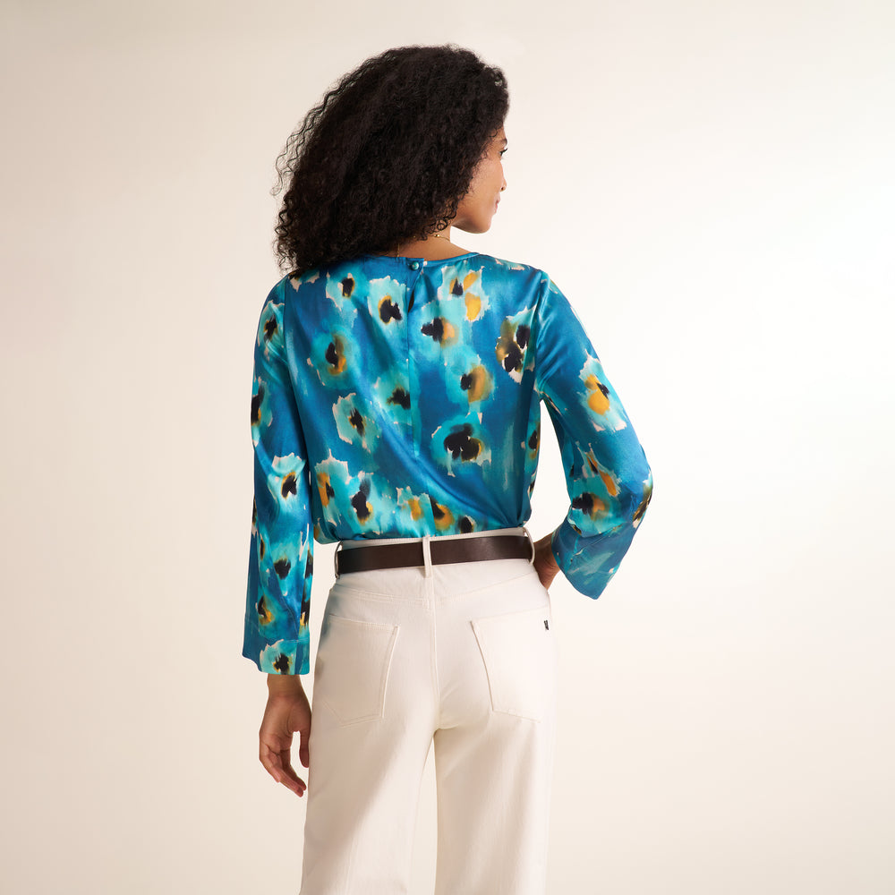 A woman shown from the back wearing a long-sleeve satin blouse with a blue, teal, black, and gold abstract pattern, paired with white trousers and a dark brown belt, against a plain off-white background.