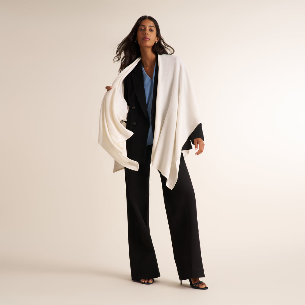 A woman models a stylish outfit featuring a black double-breasted blazer, wide-leg black trousers, a light blue top, and a draped white wrap shawl, set against a plain beige studio background.