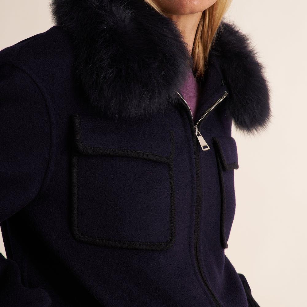 Close-up of a dark navy wool jacket with a large dark fur collar, featuring a front zipper and a large patch pocket on the chest.