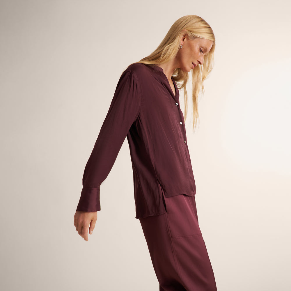 A blonde model wearing a matching burgundy long-sleeve button-up shirt and trousers set, photographed against a clean, light beige studio background.