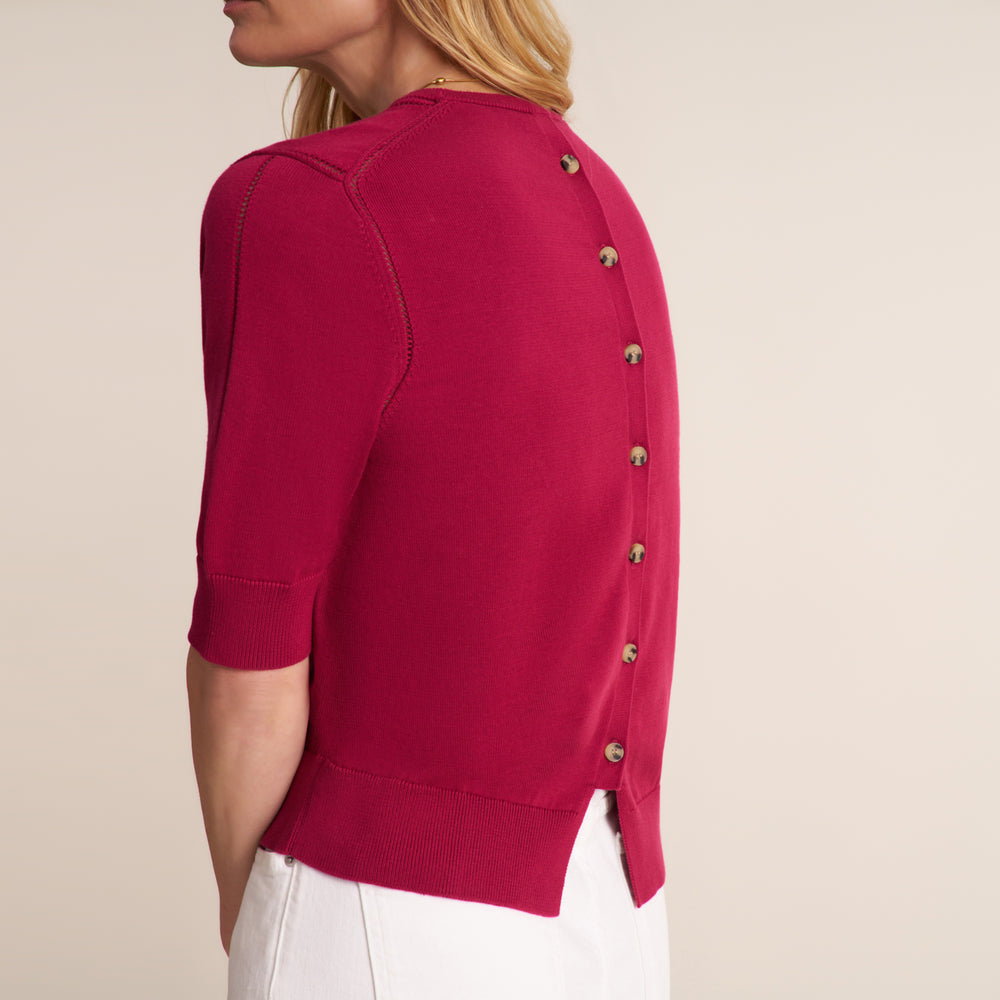 A woman wearing a bright magenta knit cardigan with half sleeves and decorative metallic buttons down the back, paired with white pants against a plain beige background.