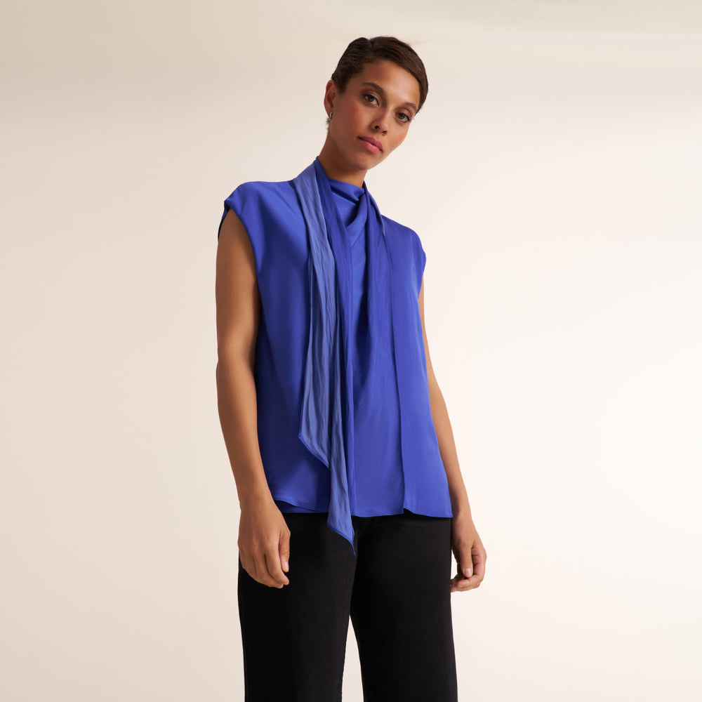 A woman modeling a sleeveless royal blue top with a draped, layered front design and black pants, photographed against a plain light beige studio background.