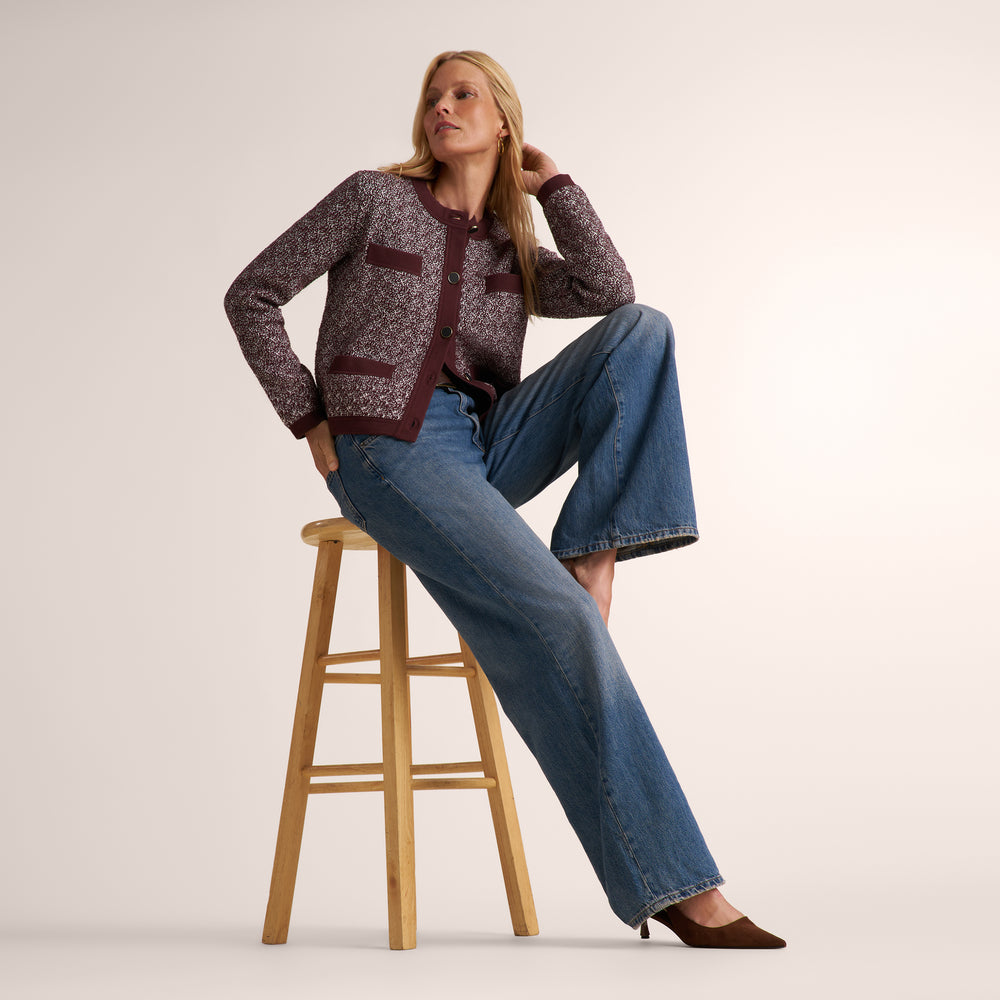 A woman models a patterned cardigan and wide-leg blue jeans while sitting on a wooden stool against a plain beige studio background.