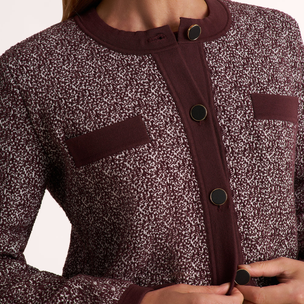 Close-up of a person wearing a textured tweed cardigan in a dark burgundy and white speckled pattern, featuring a solid burgundy placket with dark buttons and matching chest pockets.