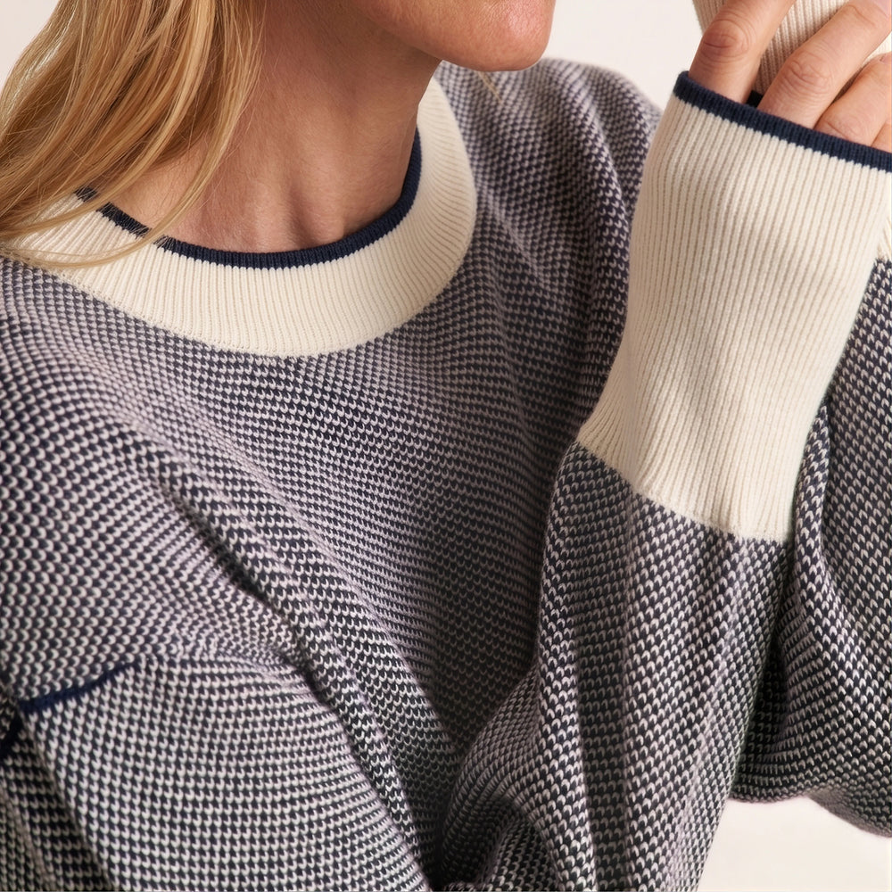 Close-up of a person wearing a textured knit sweater with a gray and white houndstooth pattern, cream ribbed collar and sleeve cuffs, and navy blue trim.