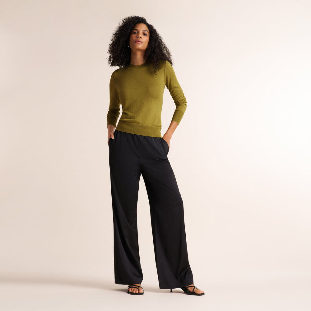 A woman modeling a long-sleeve olive green knit top and black wide-leg trousers against a plain warm off-white studio background.