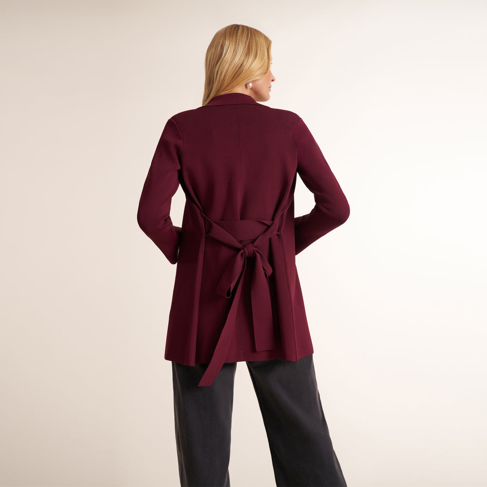 A woman shown from the back wearing a long-sleeve wrap-style cardigan in deep burgundy with a tied waist belt, paired with dark charcoal pants, against a plain light beige studio background.