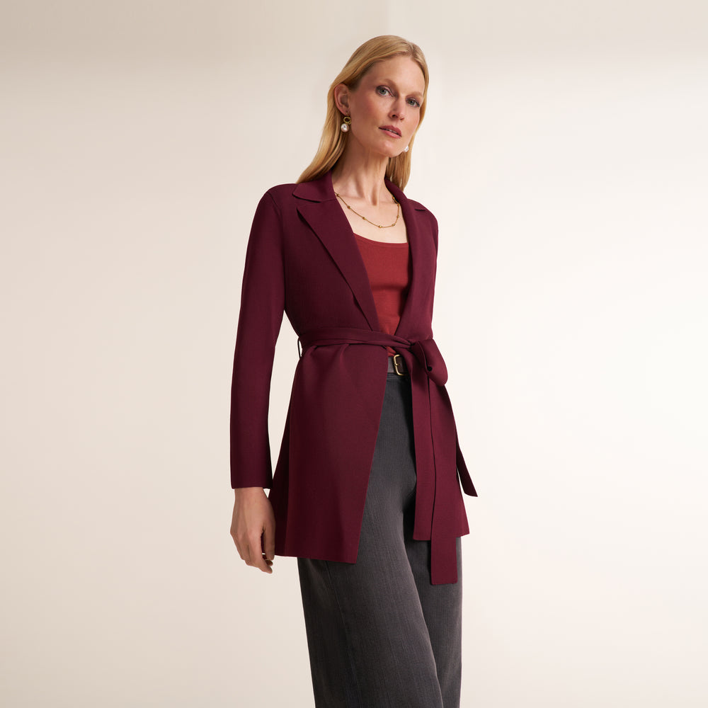 A woman modeling a long-sleeve wrap-style blazer in deep burgundy, worn over a rust-red top and dark gray trousers, against a plain light beige studio background.