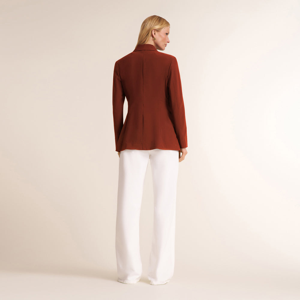 A woman shown from the back wearing a structured, long-sleeved rust-brown blazer and white wide-leg trousers, standing against a plain beige studio background.