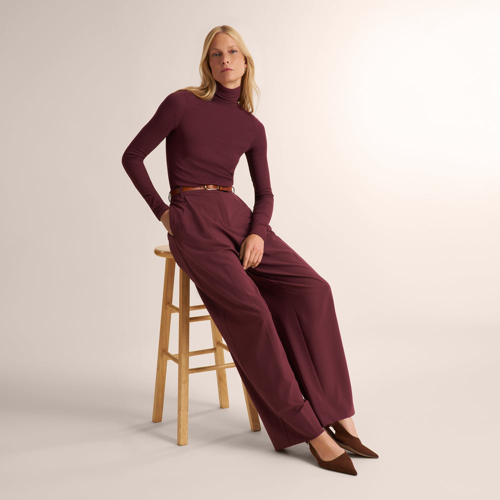 A woman models a matching burgundy turtleneck top and wide-leg trousers set with a thin brown belt, seated on a wooden stool against a warm off-white studio background.