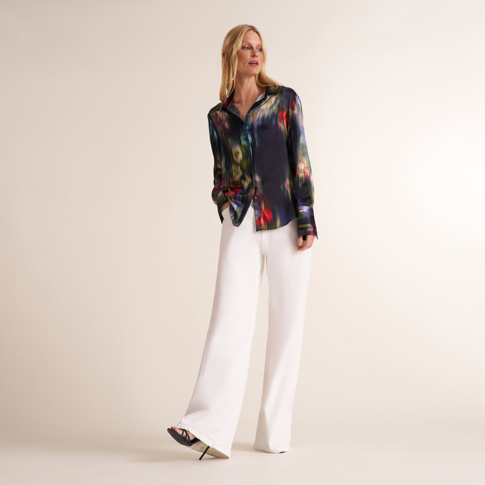 A woman modeling a long-sleeve button-up shirt with a dark abstract multicolor print, paired with white wide-leg trousers, against a plain beige studio background.