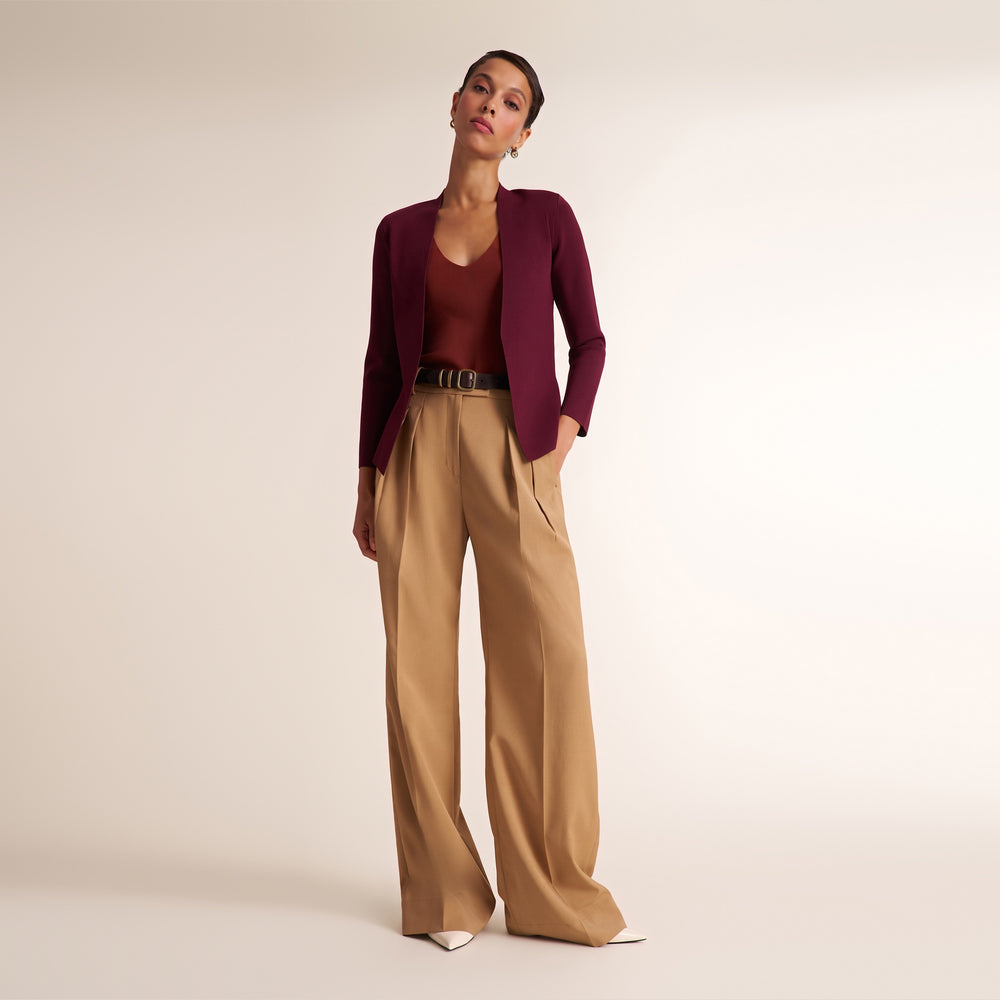 A model stands against a plain light beige studio background wearing a dark burgundy open-front cardigan over a matching burgundy top, paired with high-waisted tan wide-leg trousers and white pointed shoes.
