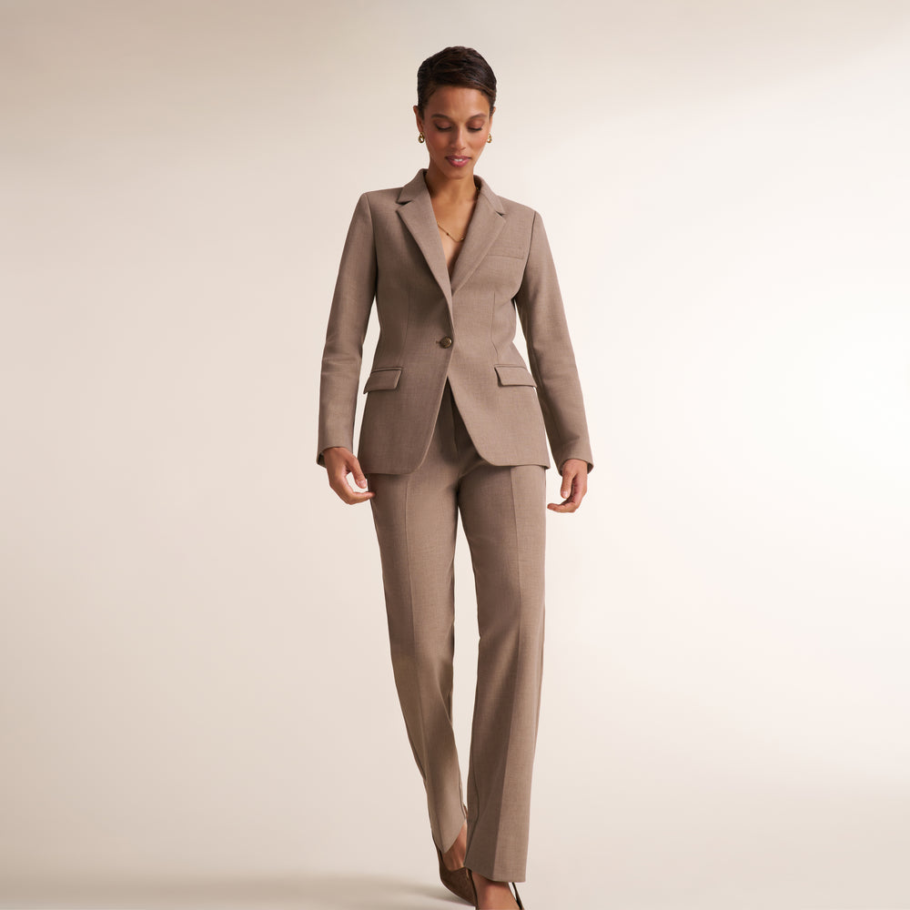 A woman modeling a tailored taupe two-piece suit with a blazer and matching trousers, standing against a plain light beige studio background.