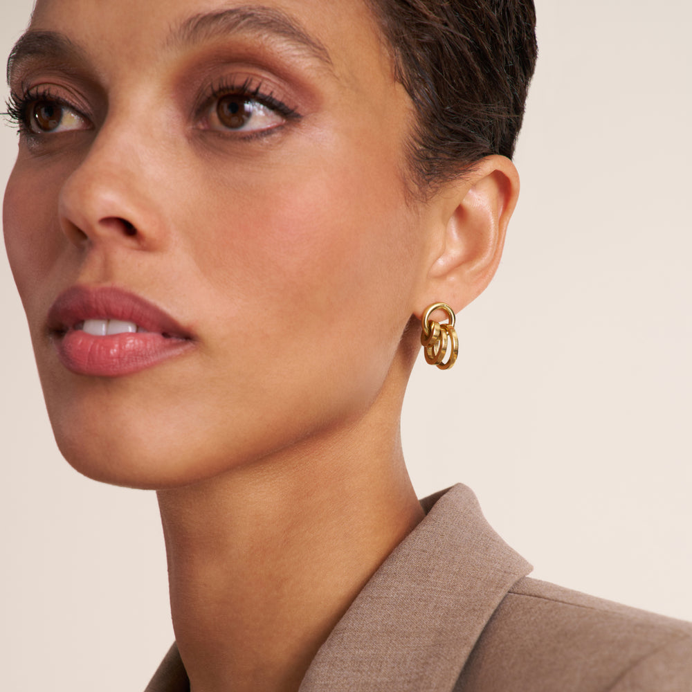 A close-up of a model wearing a gold double-ring hoop earring, shown from the side against a warm beige background.