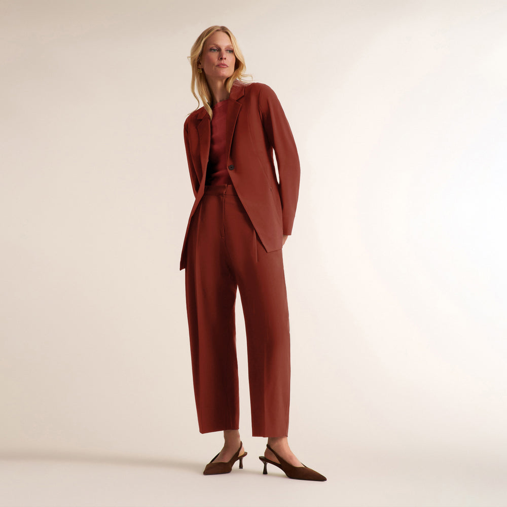 A model stands against a plain beige studio background wearing a rust-brown tailored suit set consisting of a structured blazer and matching wide-leg cropped trousers, paired with dark brown pointed-toe heels.